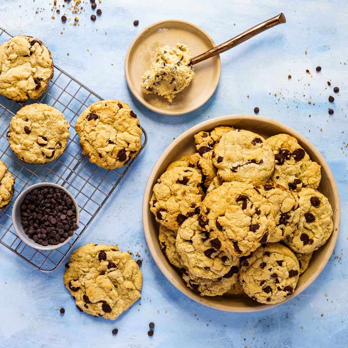 A bowl and cooling rack filled with gluten-free chocolate chip cookies, a small bowl of chocolate chips, and a plate with cookie dough and a spoon, all on a light blue surface.