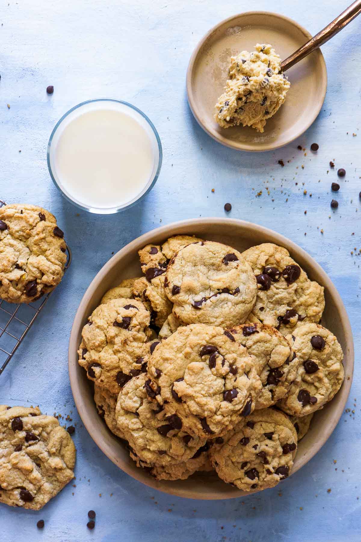 A bowl filled with gluten-free chocolate chip cookies, a glass of milk, a plate with cookie dough and a spoon, and a cooling rack with cookies sit on a light blue surface.