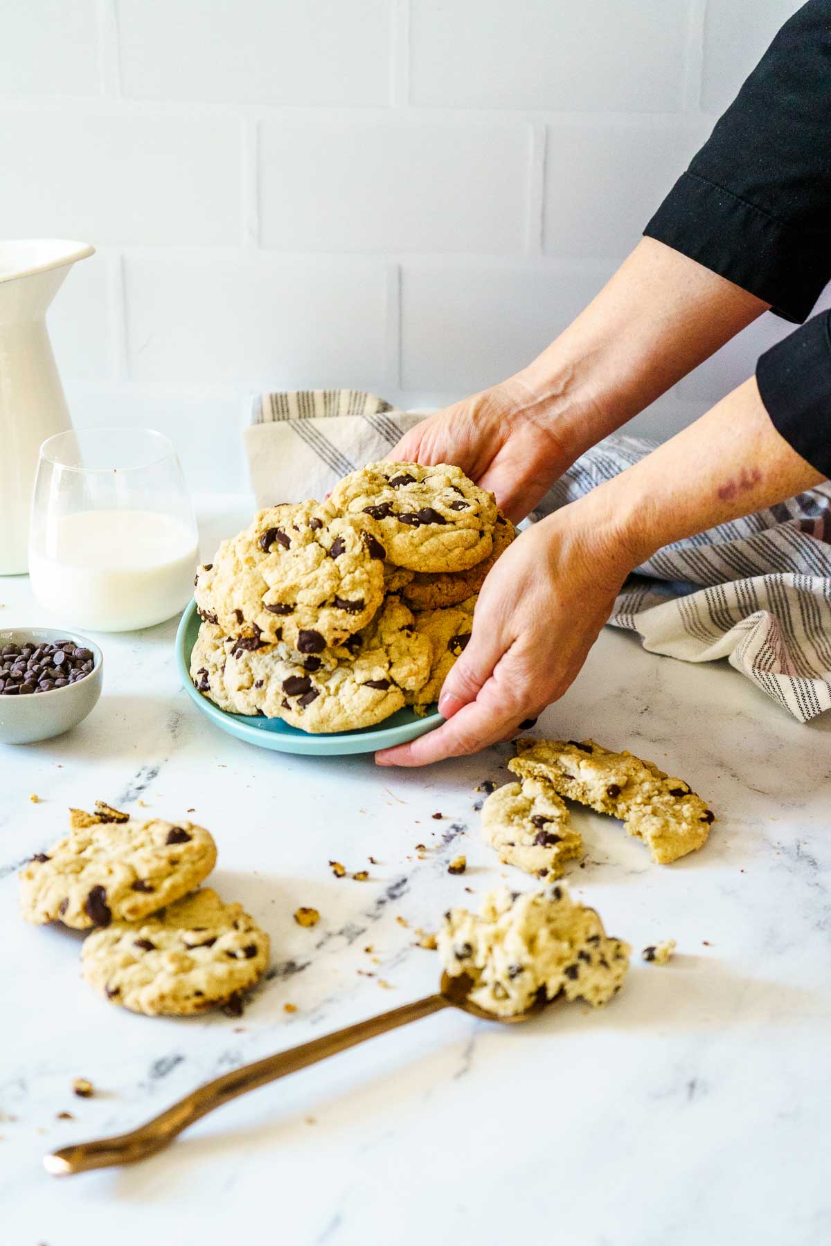A person places a plate of gluten-free chocolate chip cookies on a marble counter, with milk, chocolate chips, and a napkin nearby. Two cookies and a spoon rest on the counter.