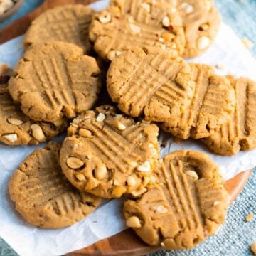 A pile of peanut butter cookies with fork marks and chopped peanuts, arranged on parchment paper atop a wooden surface.