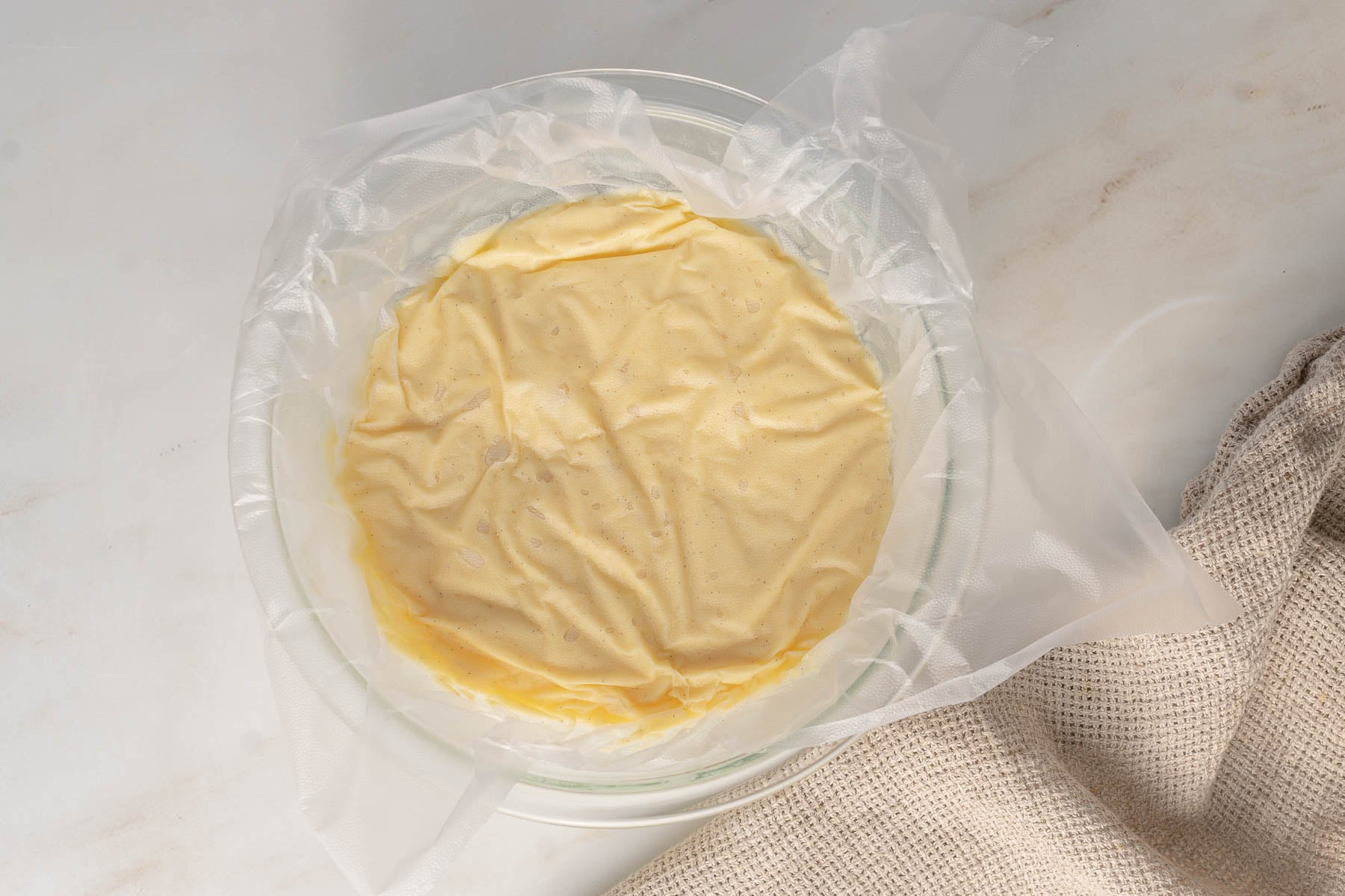 A bowl of pastry cream covered with plastic wrap rests on a light-colored surface next to a beige textured cloth.