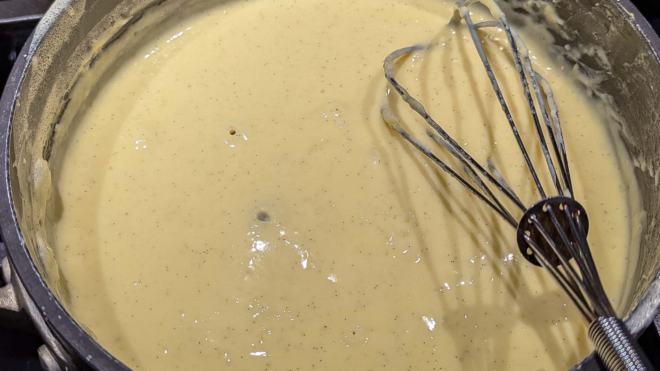 A metal whisk rests in a saucepan filled with thick, yellow pastry cream custard in the process of being mixed.