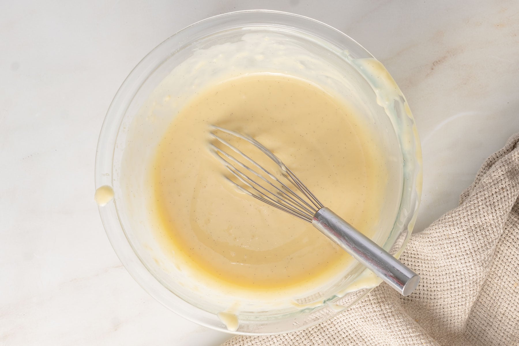 A glass bowl filled with pale yellow pastry cream batter and a metal whisk, placed on a light surface next to a beige textured cloth.