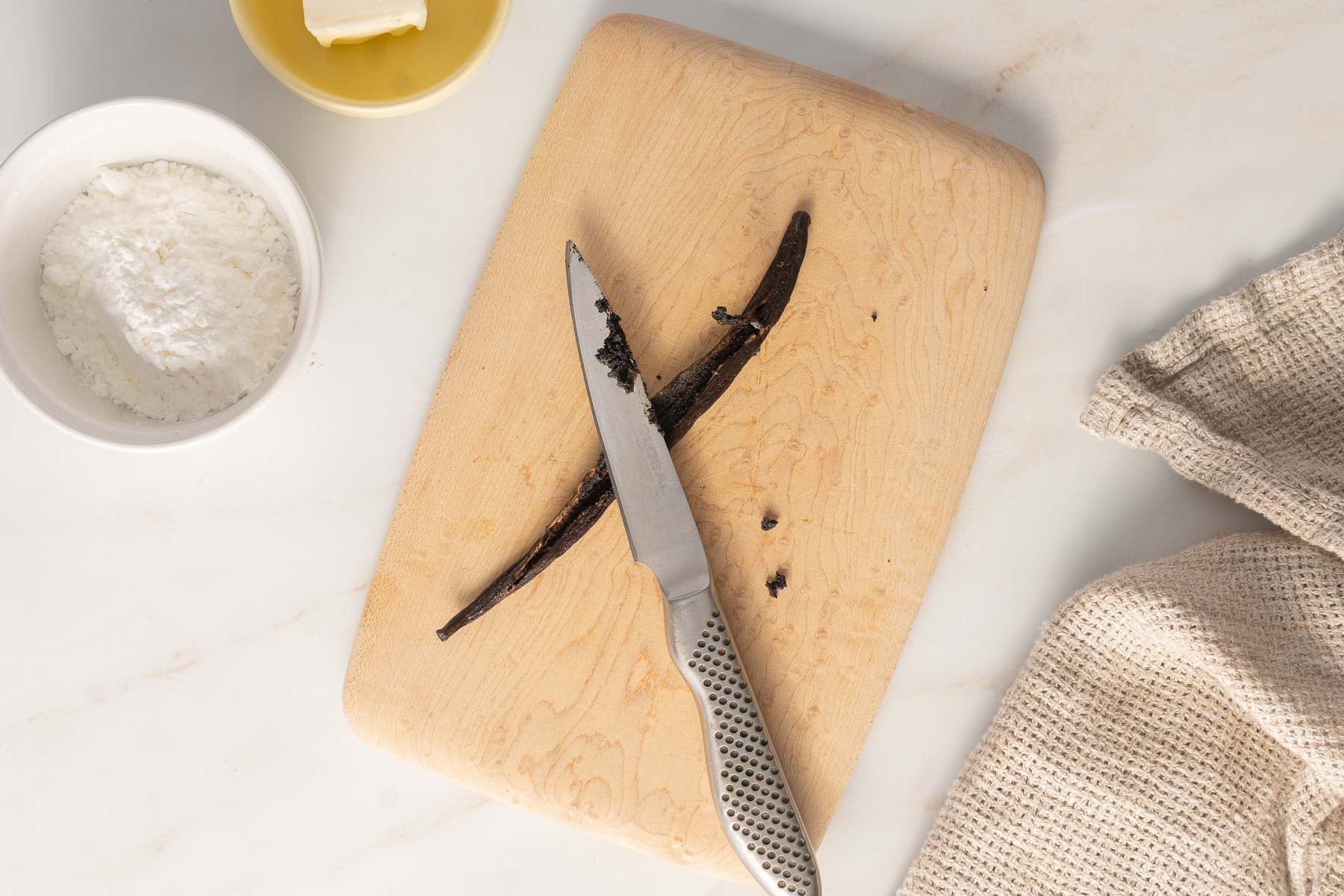 A vanilla bean pod split open on a wooden cutting board with a knife, next to a bowl of powdered sugar, a dish of butter, and a beige cloth-perfect ingredients for making rich pastry cream.