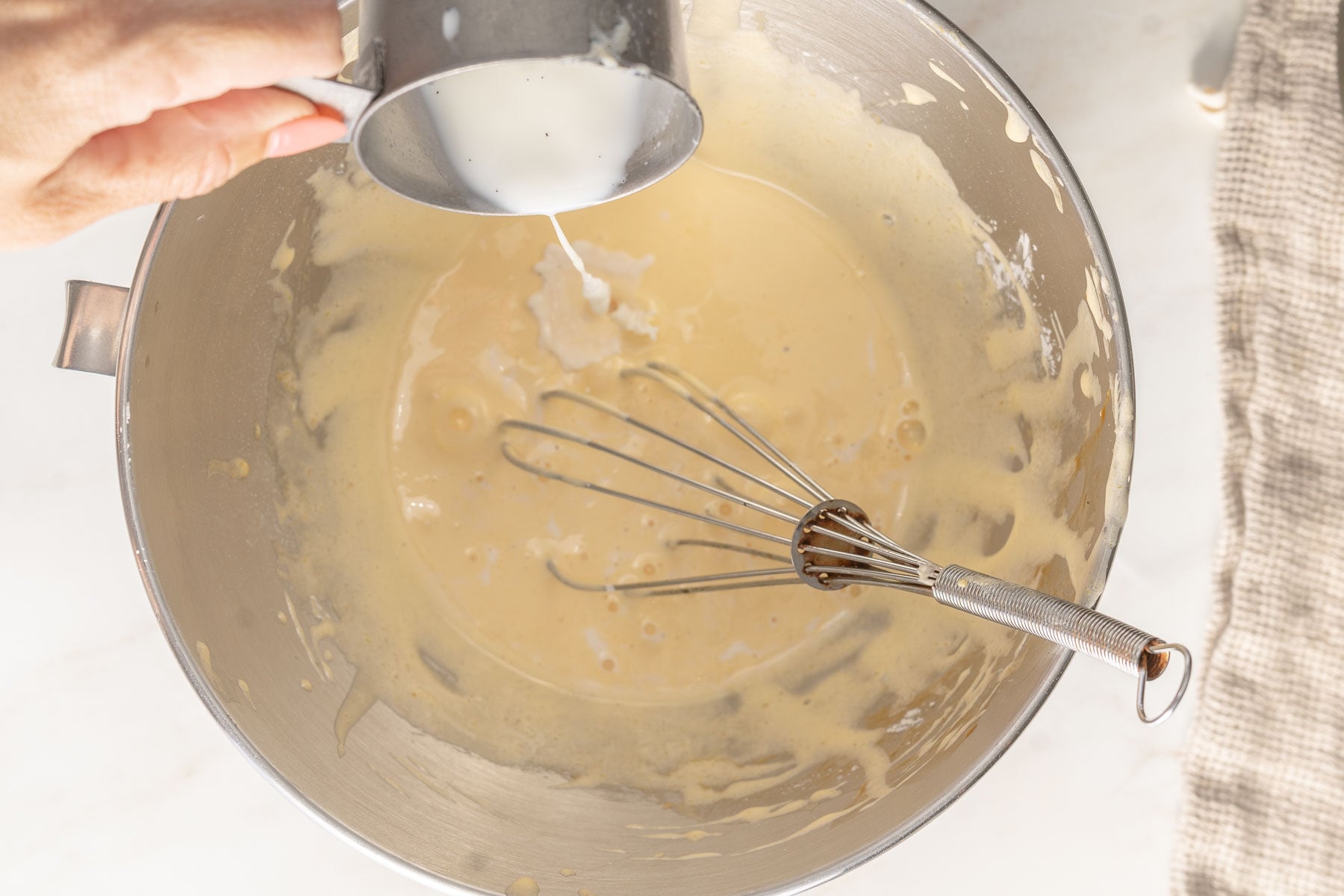A hand pours milk from a metal cup into a mixing bowl containing pastry cream batter, with a metal whisk resting inside the bowl.