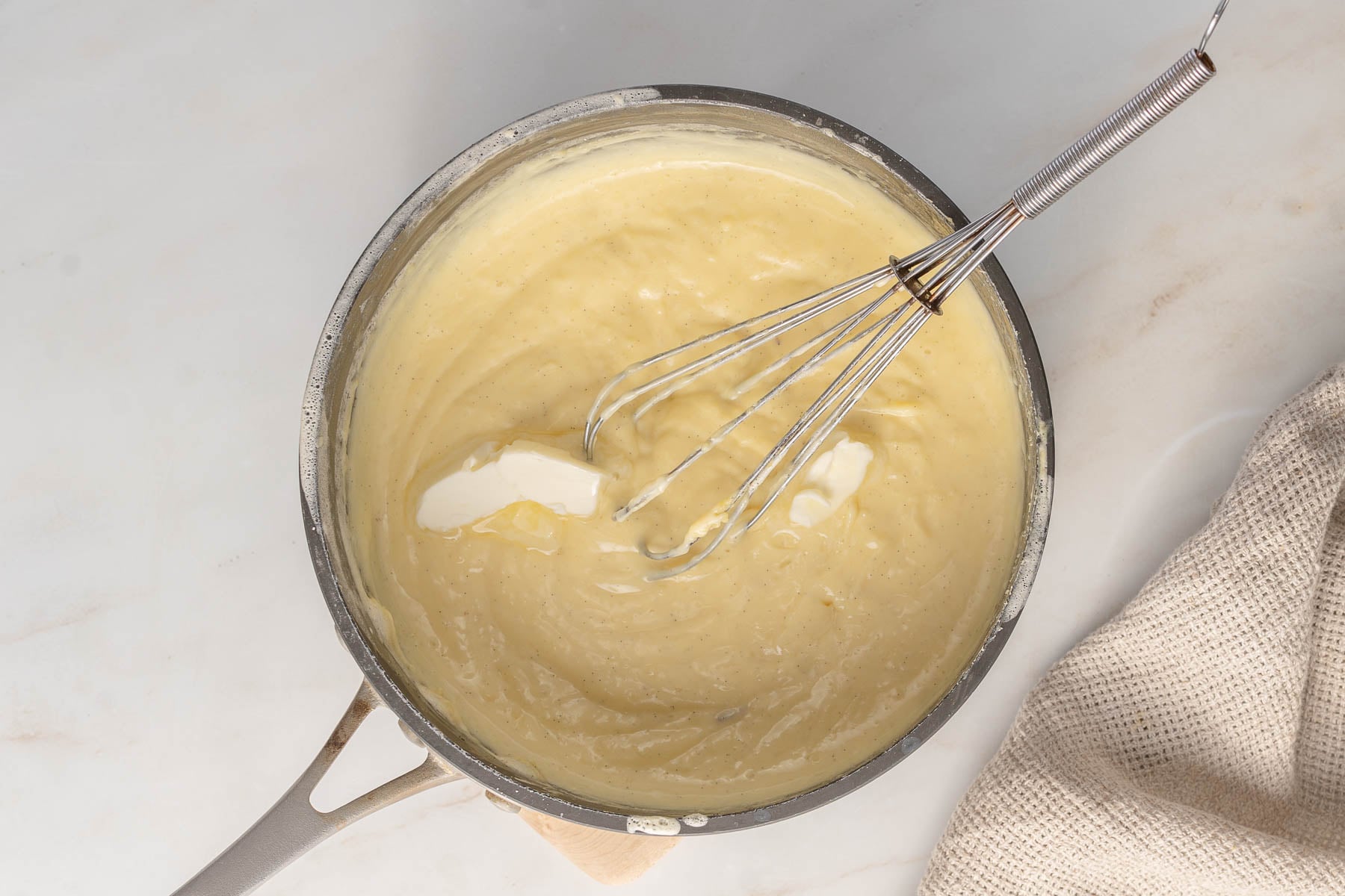 A saucepan filled with thick, creamy pastry cream being whisked, with a pat of butter melting on top; a beige textured cloth is beside the pan.