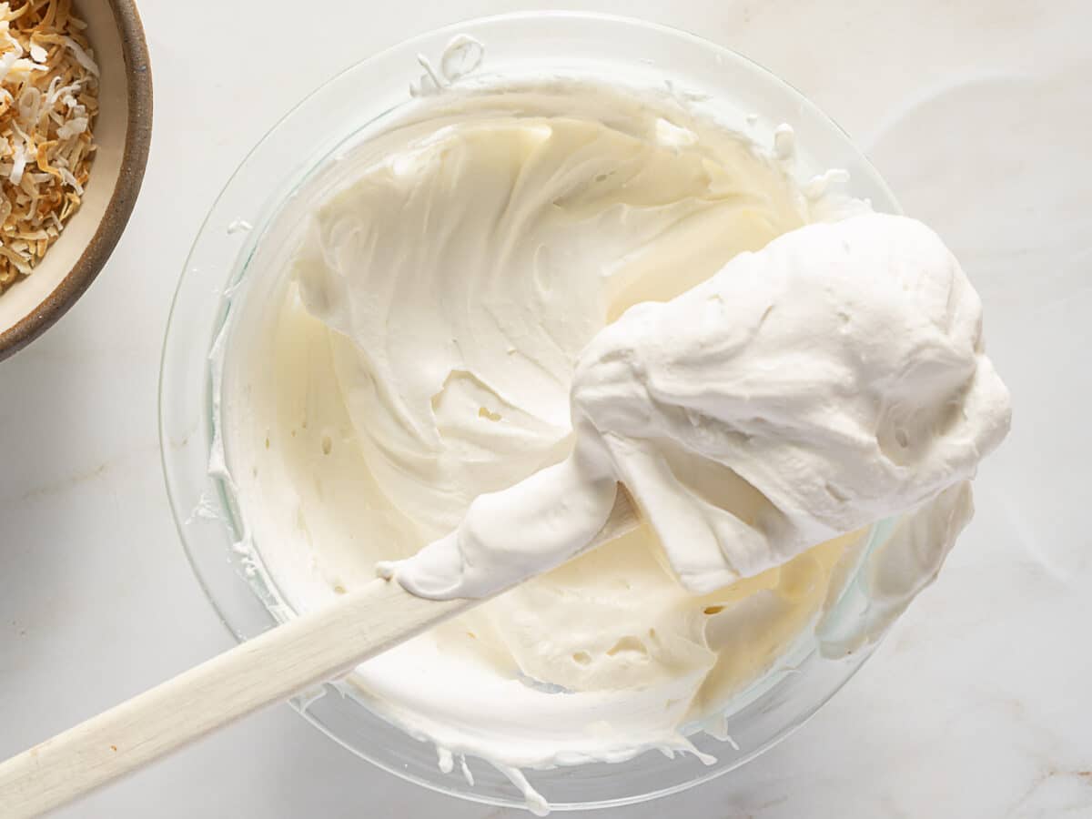 A glass bowl filled with whipped cream for a Coconut Cream Pie is being stirred with a white spatula, next to a small bowl of toasted coconut flakes—perfect for assembling a delicious Gluten Free dessert.