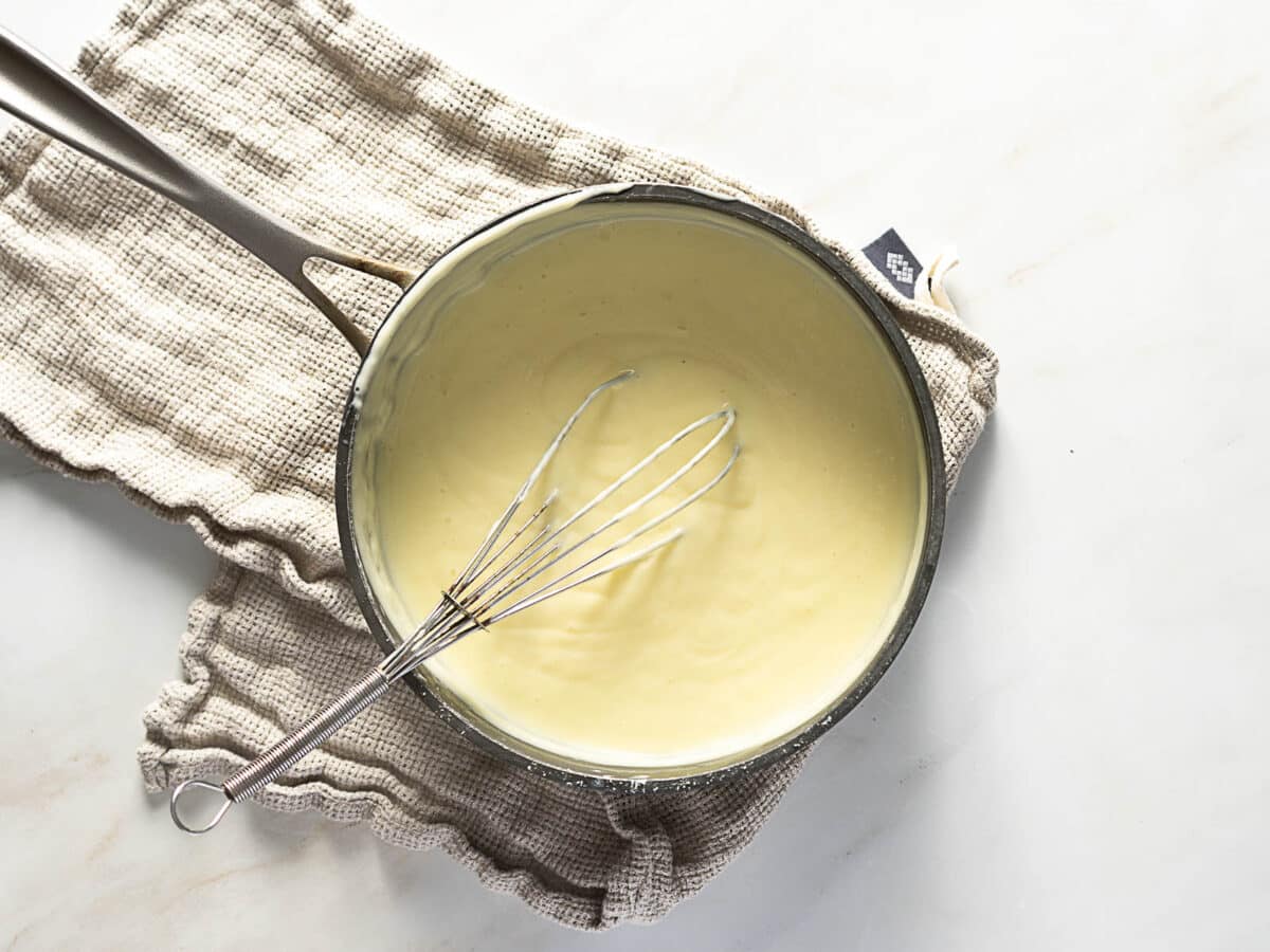 A saucepan of creamy white sauce with a whisk inside sits on a folded beige kitchen towel on a white surface.