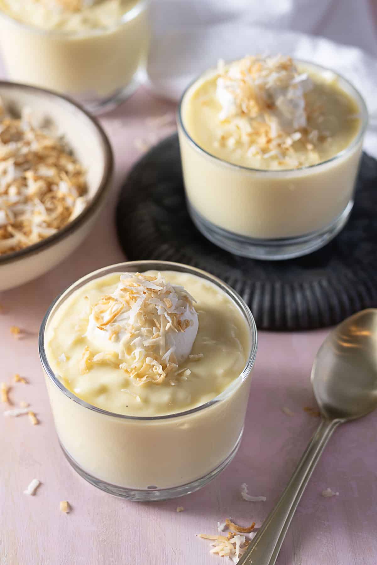 Three glass cups of creamy pudding topped with whipped cream and toasted coconut flakes, with a bowl of coconut flakes and a spoon nearby.