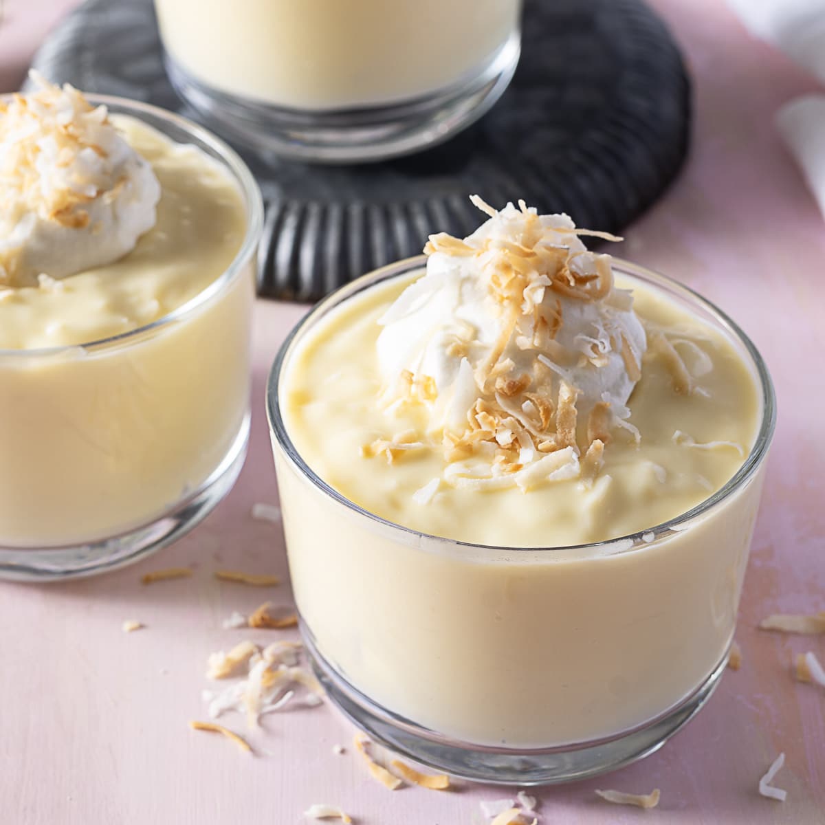 A glass cup filled with creamy pudding, topped with whipped cream and toasted coconut flakes; another similar dessert is in the background.