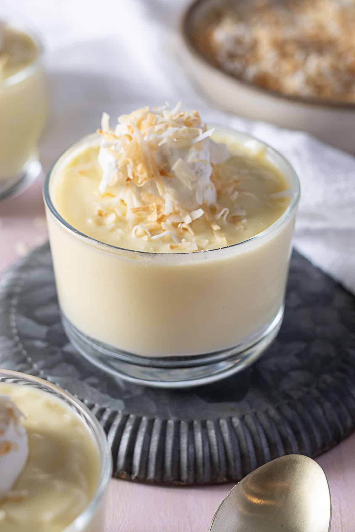 A glass cup filled with creamy pudding, topped with whipped cream and toasted coconut flakes, placed on a textured gray coaster.