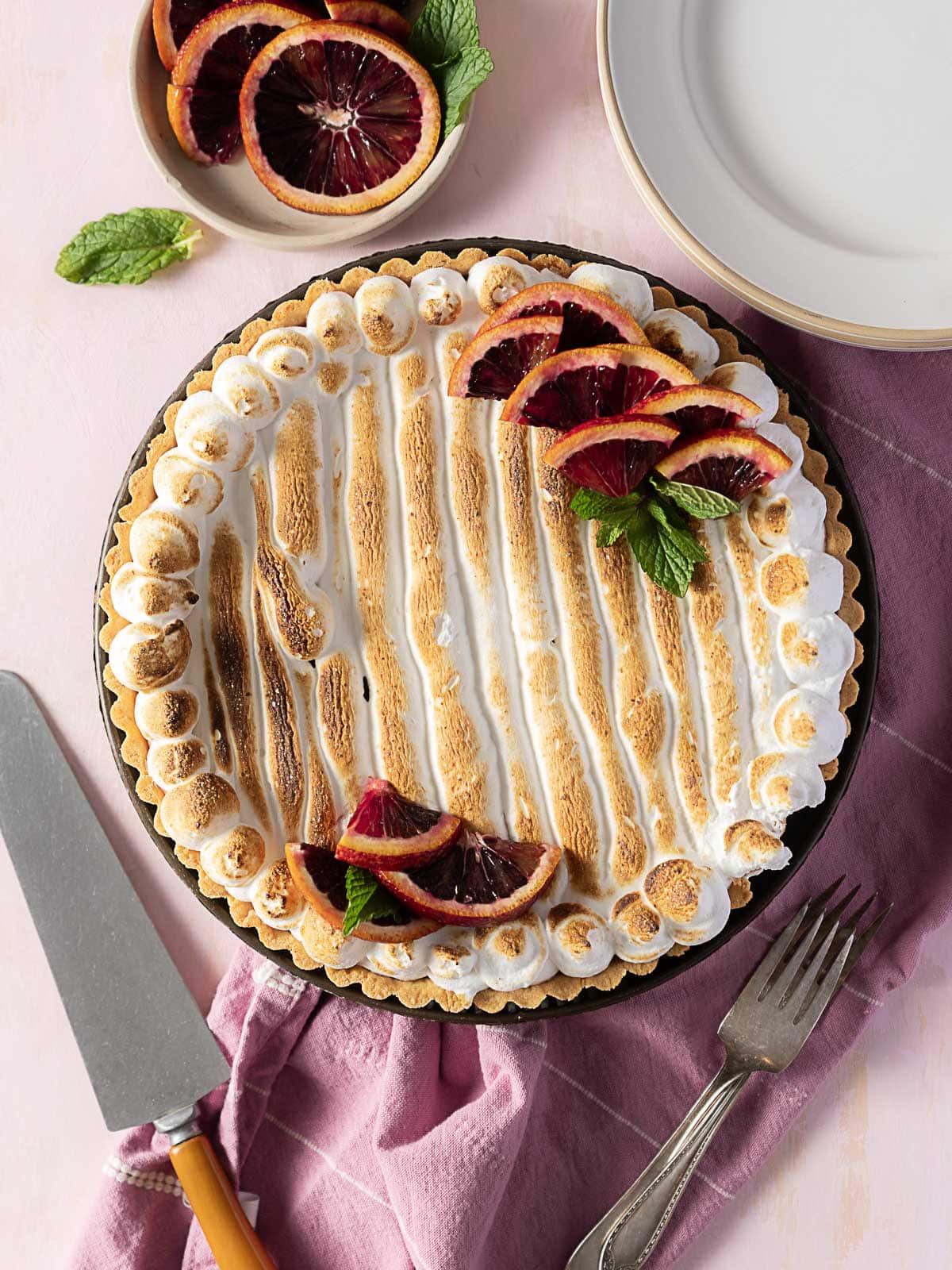A toasted meringue pie topped with blood orange slices and mint leaves, placed on a pink cloth next to serving plates, a knife, and a fork.