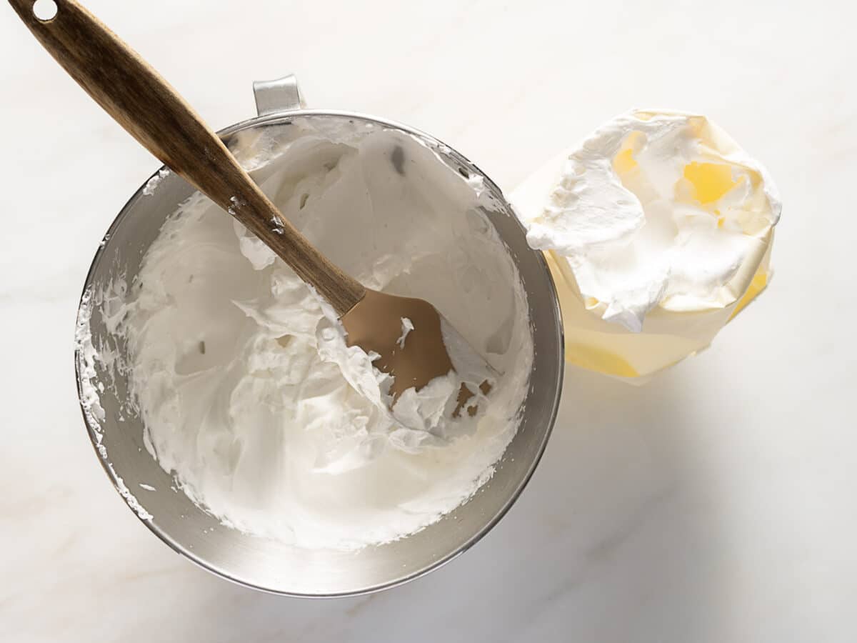 A mixing bowl with whipped cream and a spatula next to a piping bag filled with whipped cream on a light surface.