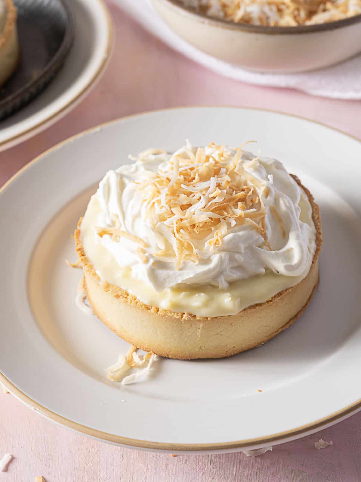 A gluten free coconut cream pie tart topped with whipped cream and toasted shredded coconut sits on a white plate.