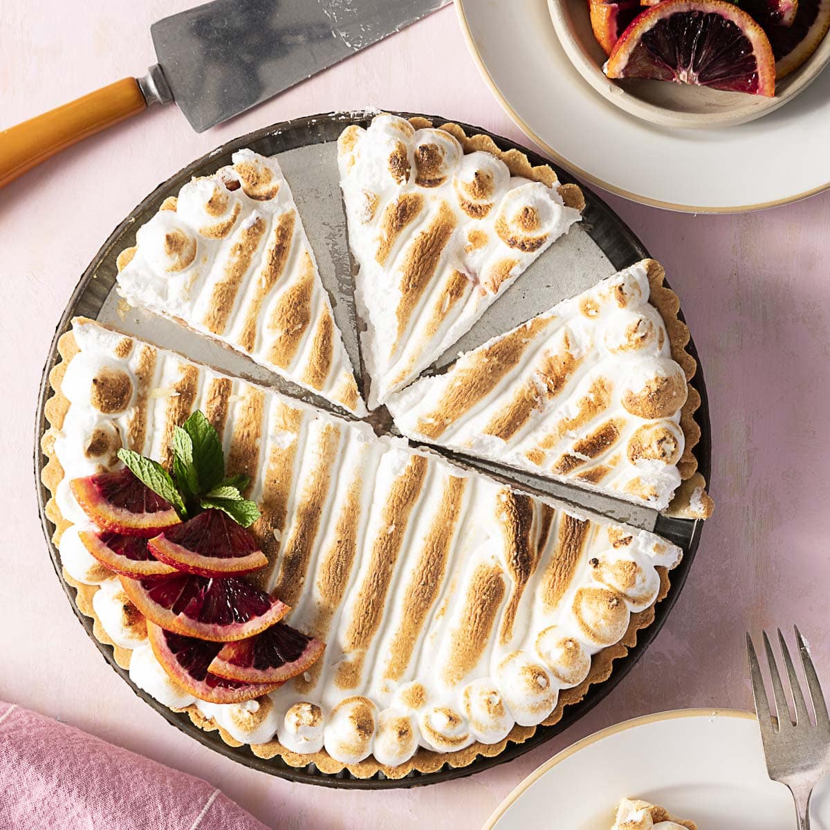 A tart topped with browned meringue and garnished with blood orange slices and mint is being sliced on a pink surface.