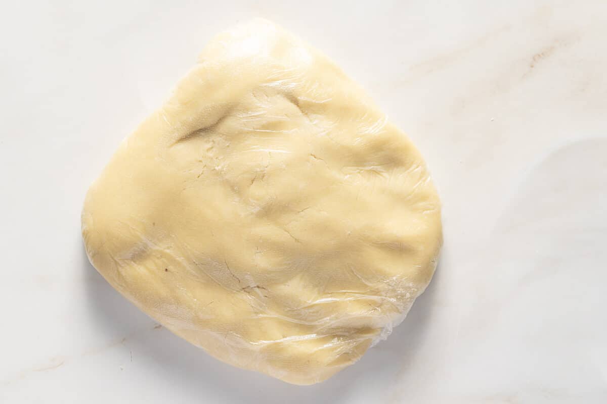 A mound of pale Sweet Tart Dough, or Pâte Sucrée, wrapped in plastic wrap rests on a light-colored marble surface.