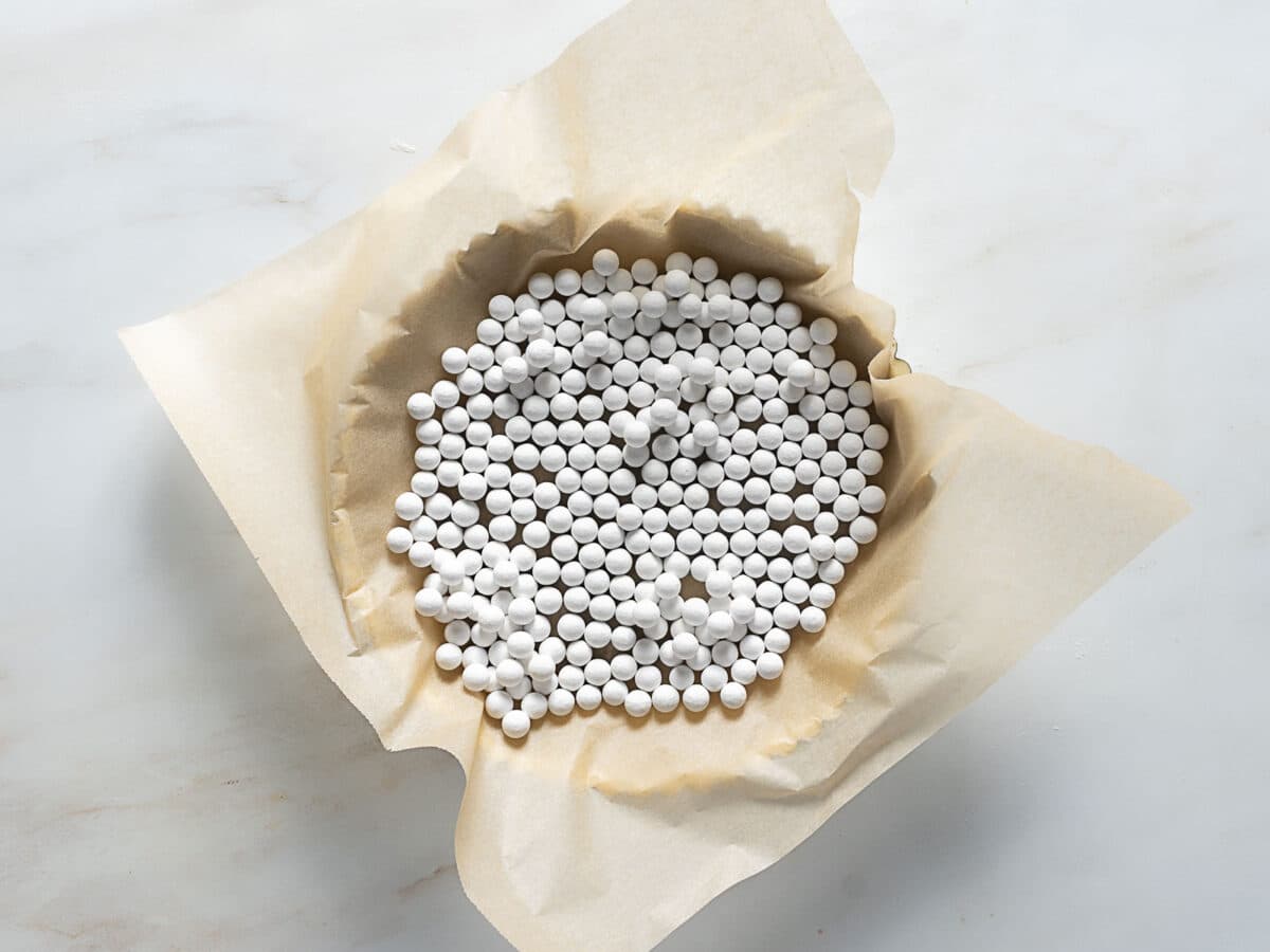 A pie dish lined with parchment paper is filled with ceramic pie weights, ready for blind baking a buttery pâte sucrée on a white surface.