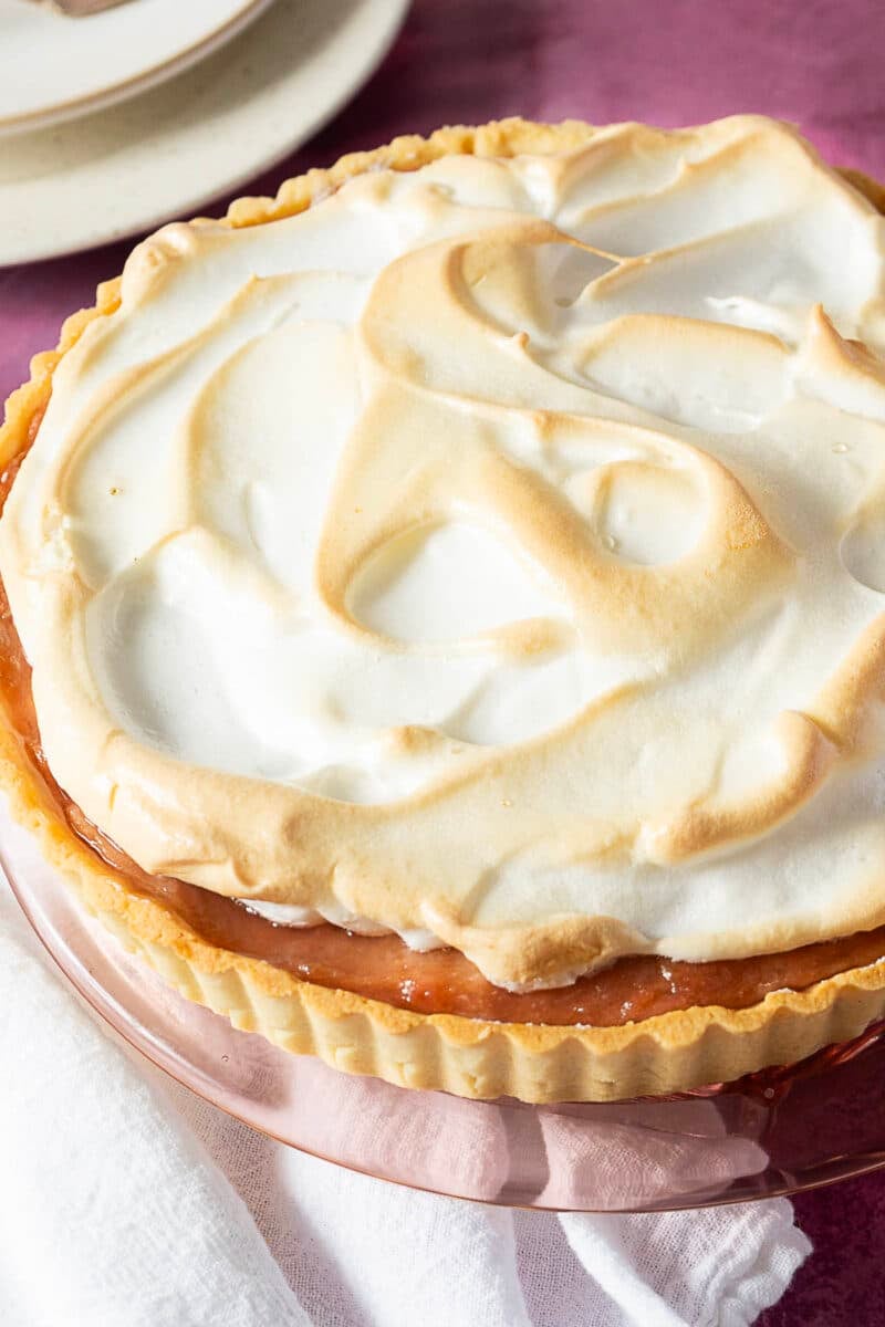 A baked pie with a golden-brown meringue topping sits on a glass plate, with part of a white cloth napkin visible in the foreground.