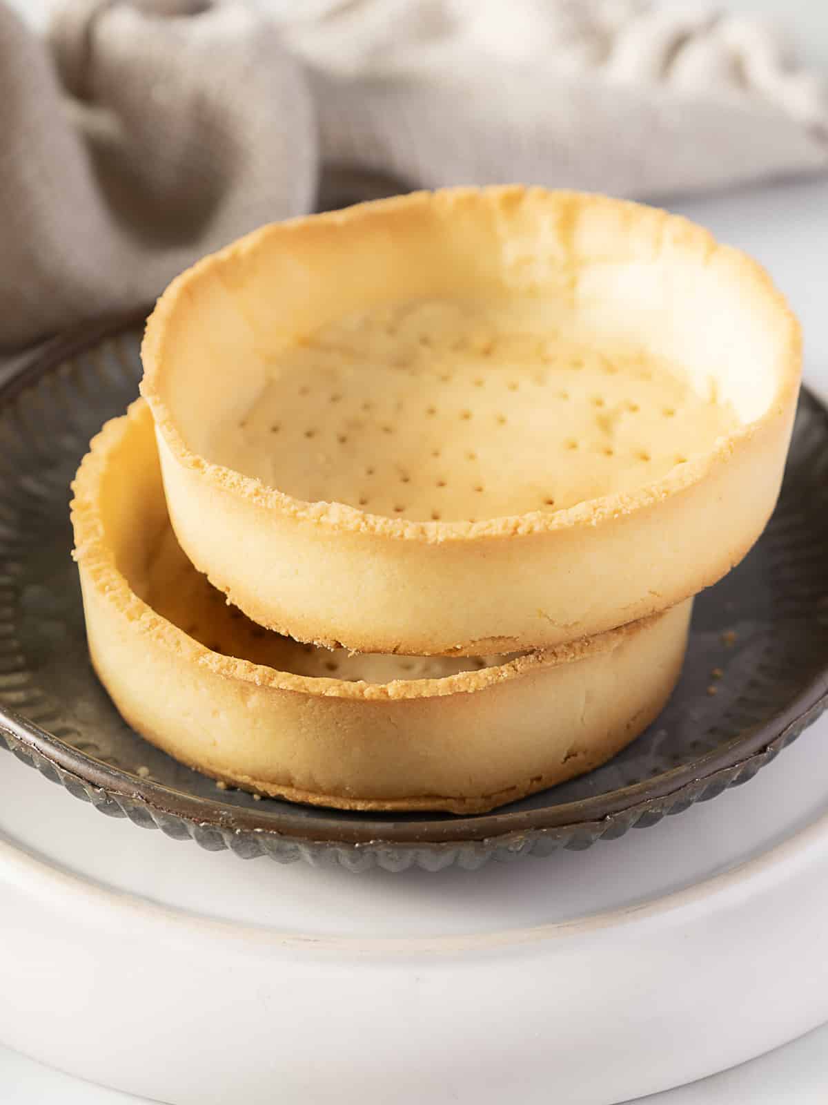 Two empty pâte sucrée tart shells are stacked on a dark plate, resting on a white surface with a textured fabric in the background.