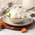 A bowl of whipped cream garnished with a mint leaf sits on a plate with a spoon, surrounded by strawberries on a wooden board.