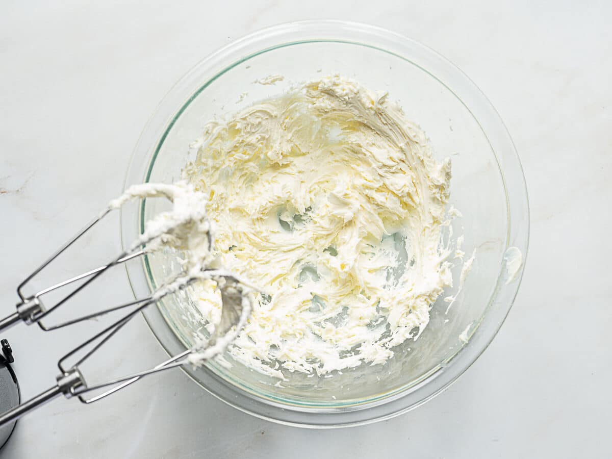 A glass bowl containing whipped cream cheese sits on a white surface next to a hand mixer with cream cheese residue on the beaters.