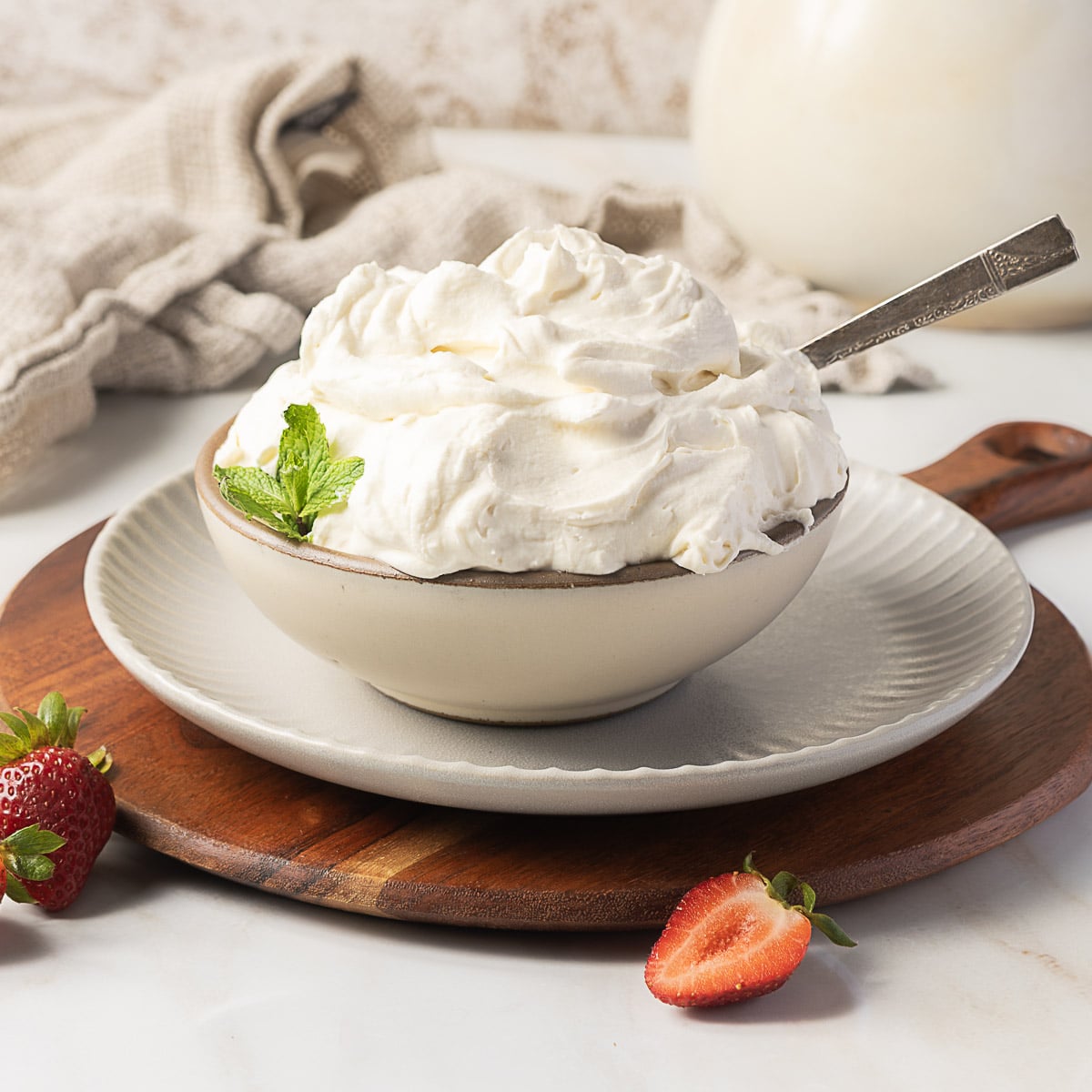 A bowl of whipped cream garnished with a mint leaf sits on a plate with a spoon, surrounded by strawberries on a wooden board.