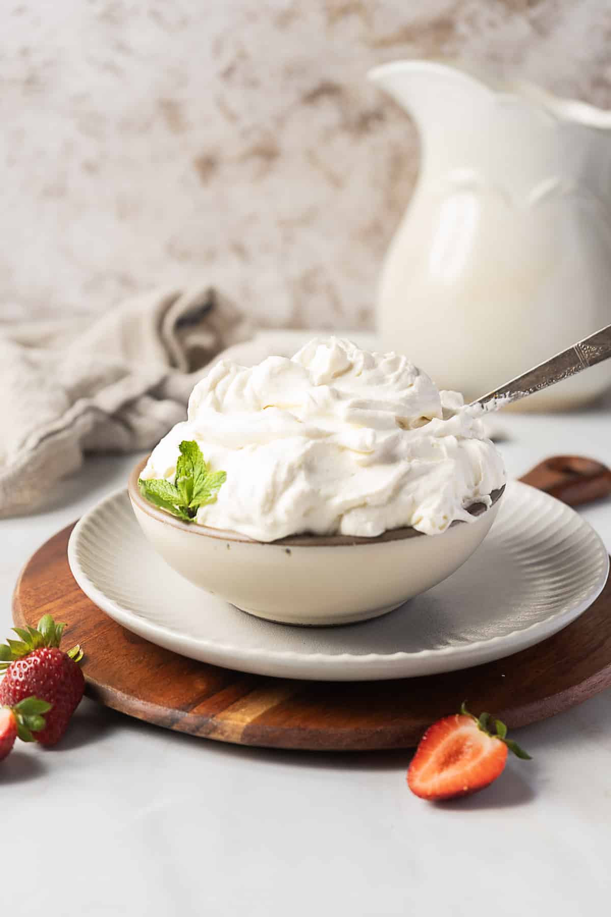 A bowl of whipped cream garnished with a mint leaf, with a spoon inside, placed on a plate and wooden board, surrounded by fresh strawberries.
