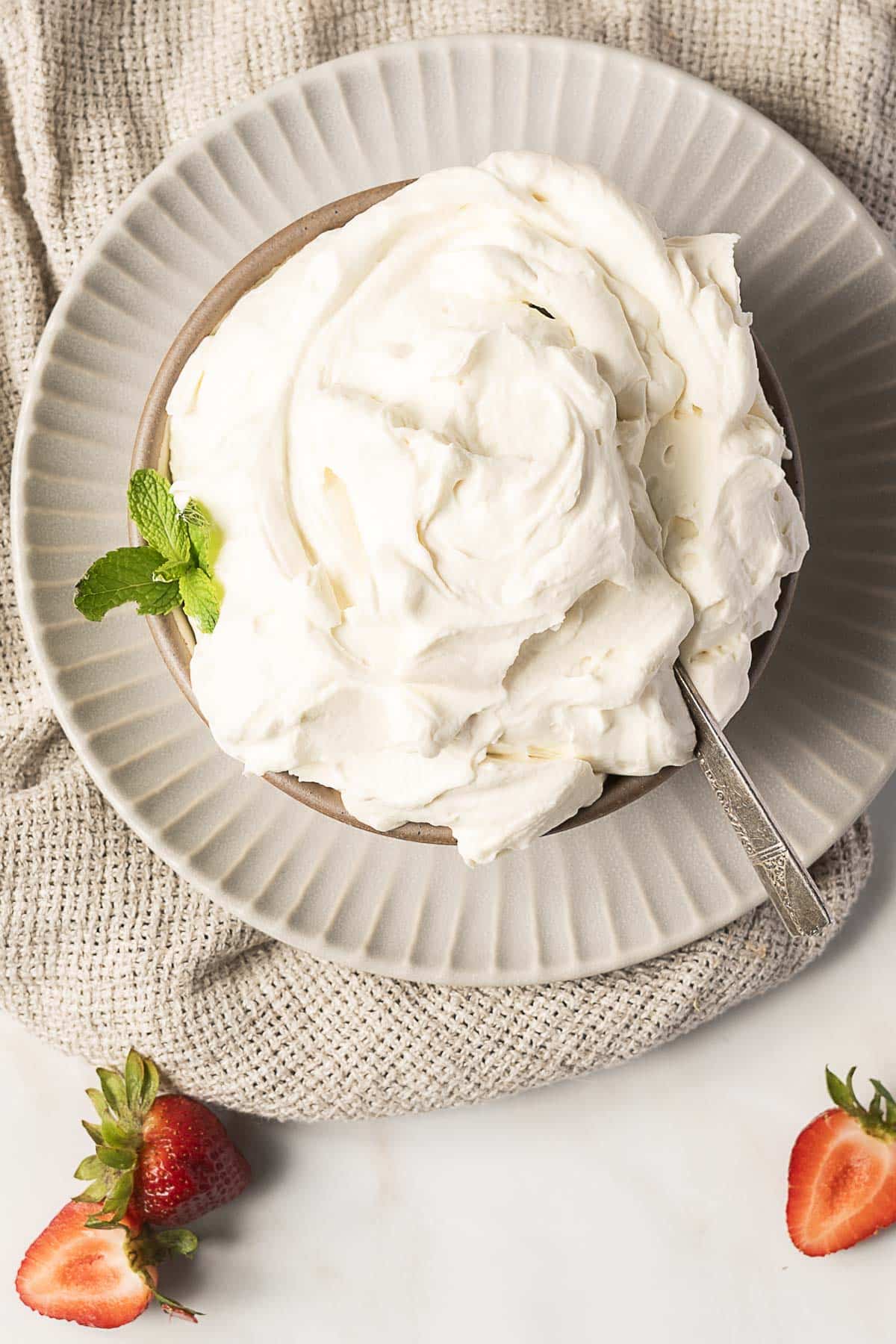 A bowl of whipped cream with a spoon, garnished with a mint sprig, placed on a plate with a textured cloth underneath and strawberries nearby.
