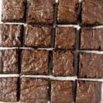A grid of sixteen evenly cut chocolate brownies arranged on a white surface, with a sprinkle of sea salt on top.
