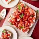 A square cake topped with whipped cream, sliced strawberries, and mint leaves on parchment paper, with plates and a serving utensil nearby.