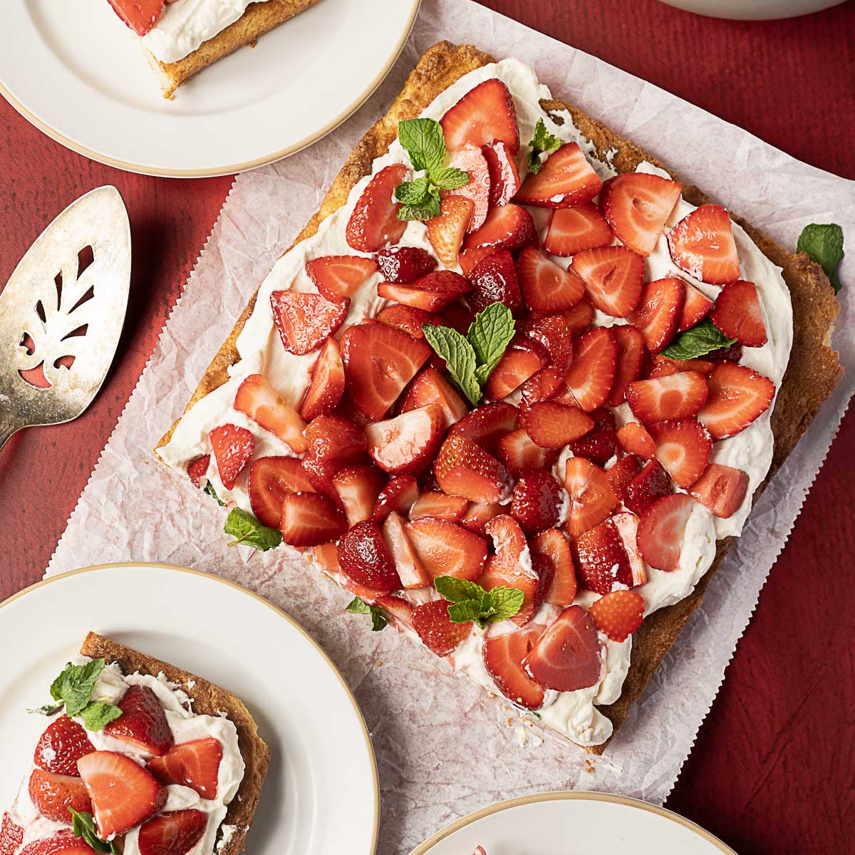 A square cake topped with whipped cream, sliced strawberries, and mint leaves on parchment paper, with plates and a serving utensil nearby.