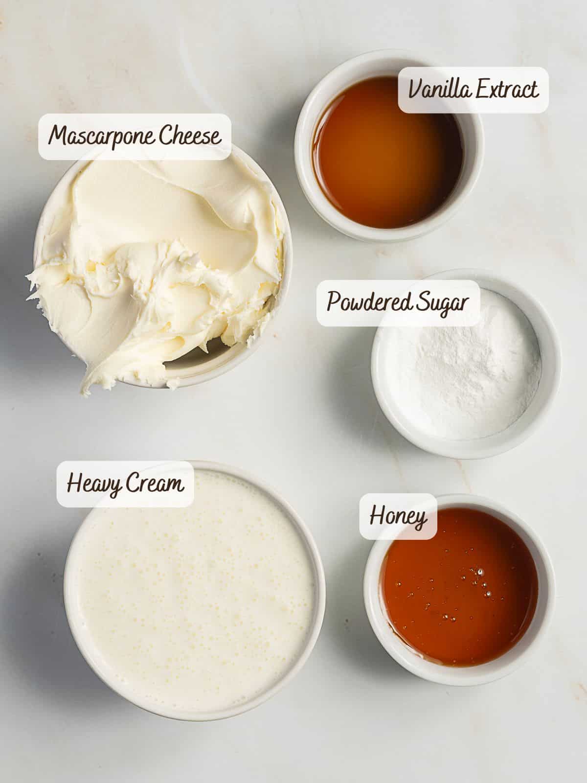 Five bowls on a white surface contain ingredients for mascarpone whipped cream: mascarpone cheese, heavy cream, vanilla extract, powdered sugar, and honey