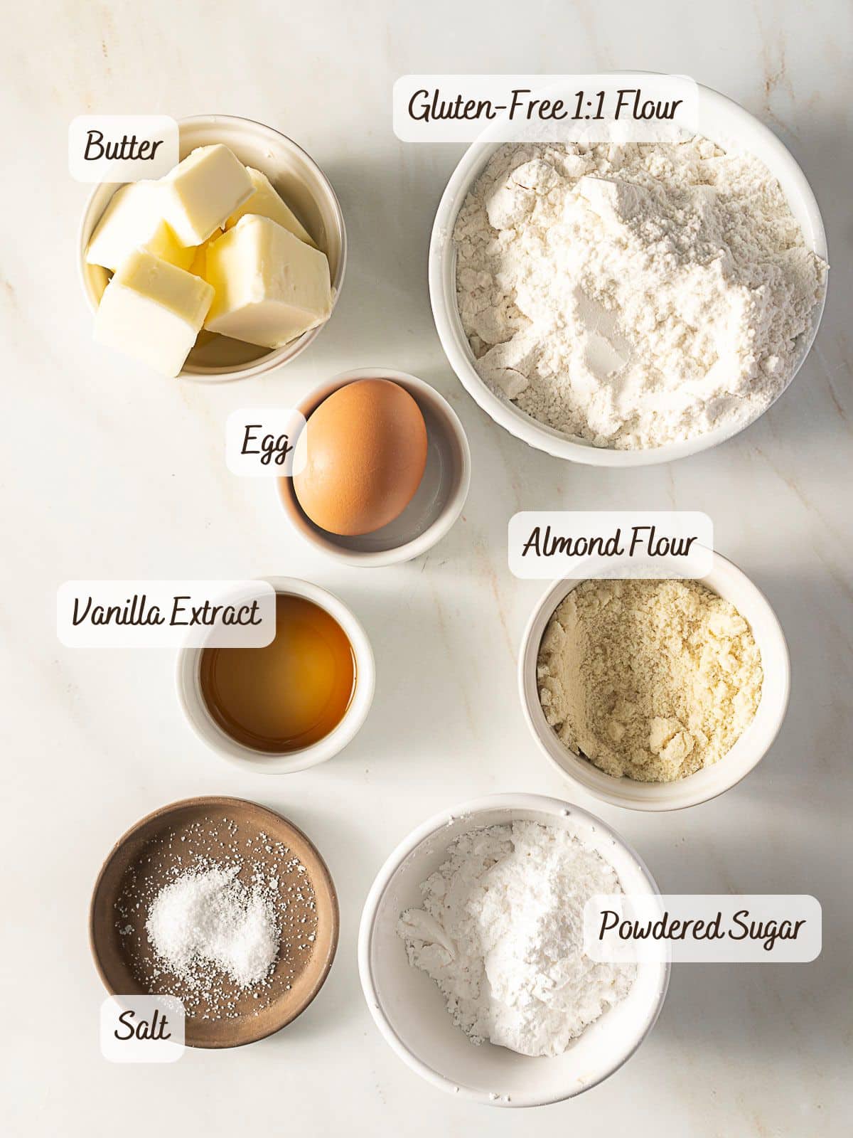 Bowls containing cubed butter, a whole egg, gluten-free 1:1 flour, almond flour, powdered sugar, salt, and vanilla extract arranged on a white surface—perfect ingredients for making gluten free pate sucree.