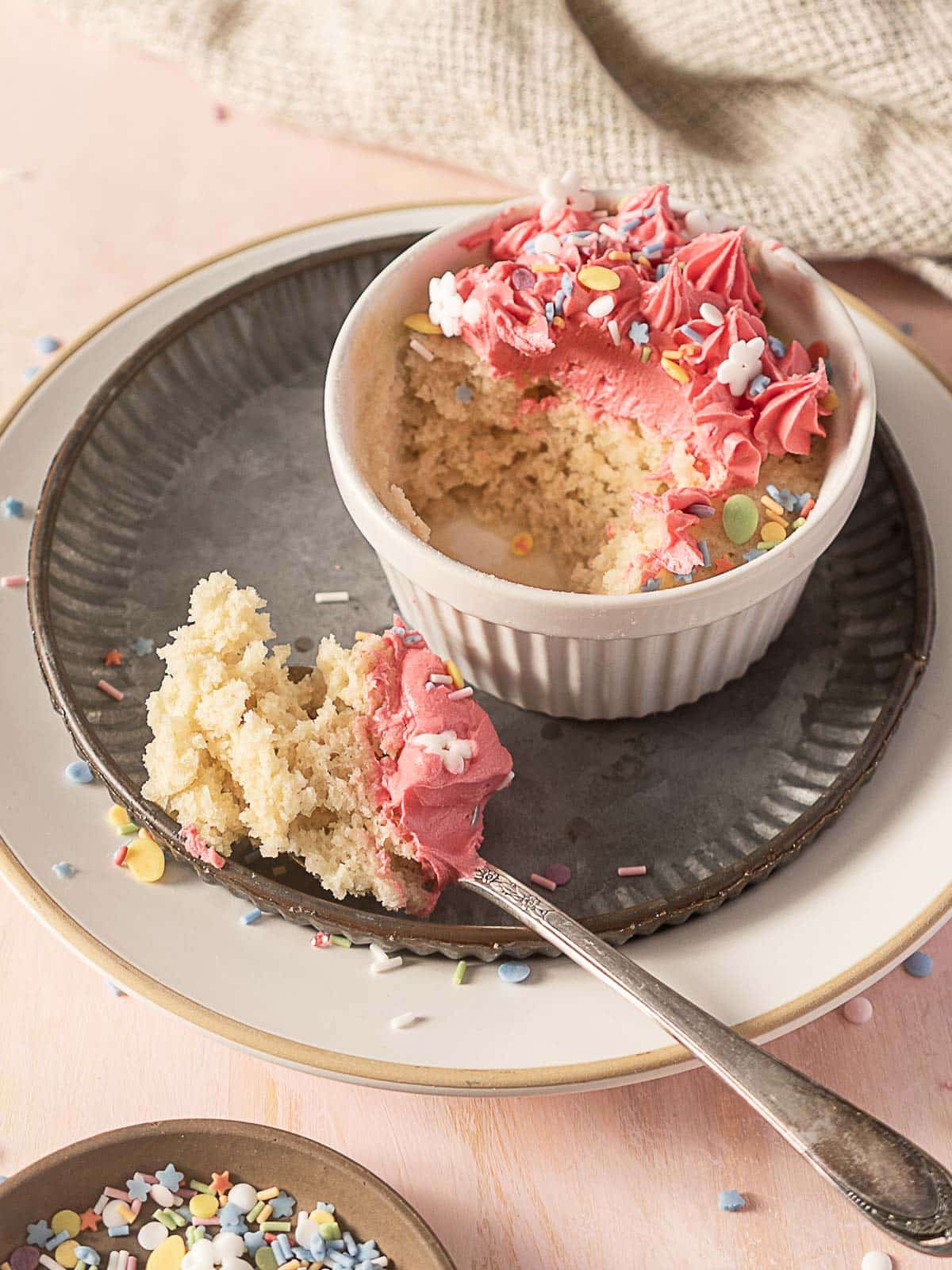 A small cake with pink frosting and colorful sprinkles in a white ramekin, with a spoonful of cake on a plate beside it.