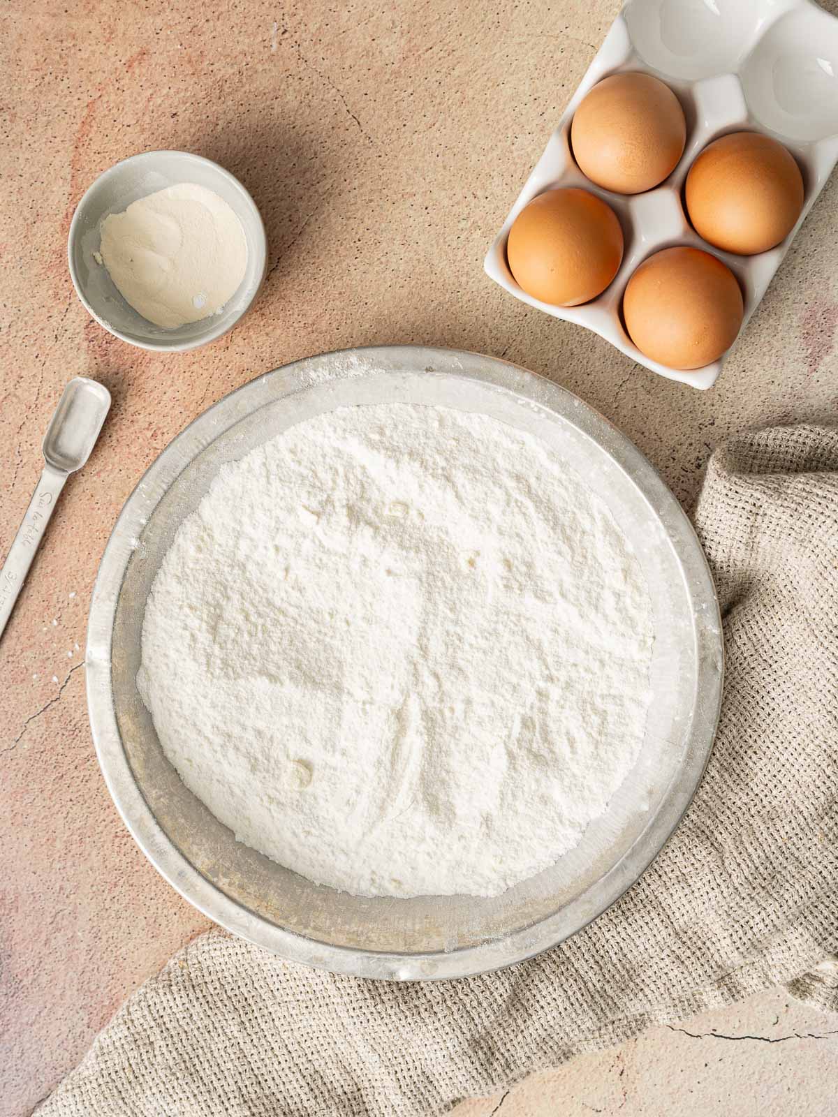 A metal bowl filled with flour, a small bowl of white powder, a measuring spoon, four brown eggs in a carton, and a beige cloth on a textured surface.
