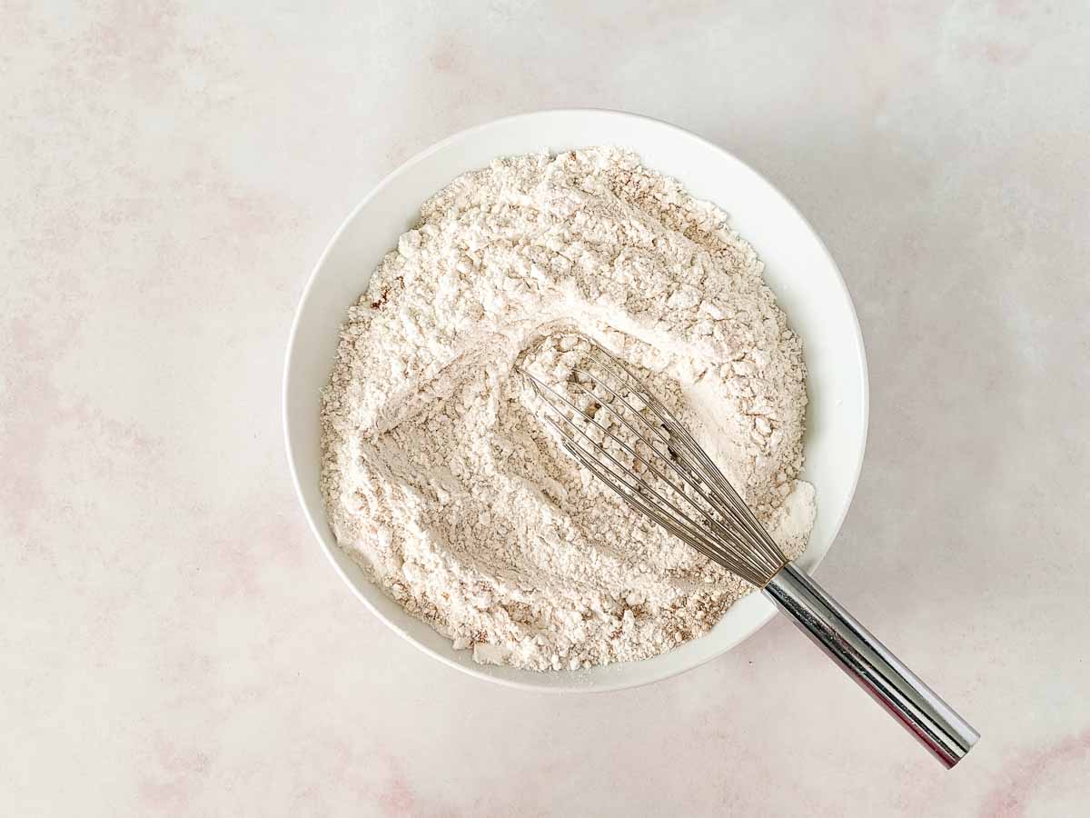 A metal whisk rests in a white bowl filled with flour mixture on a light-colored surface.