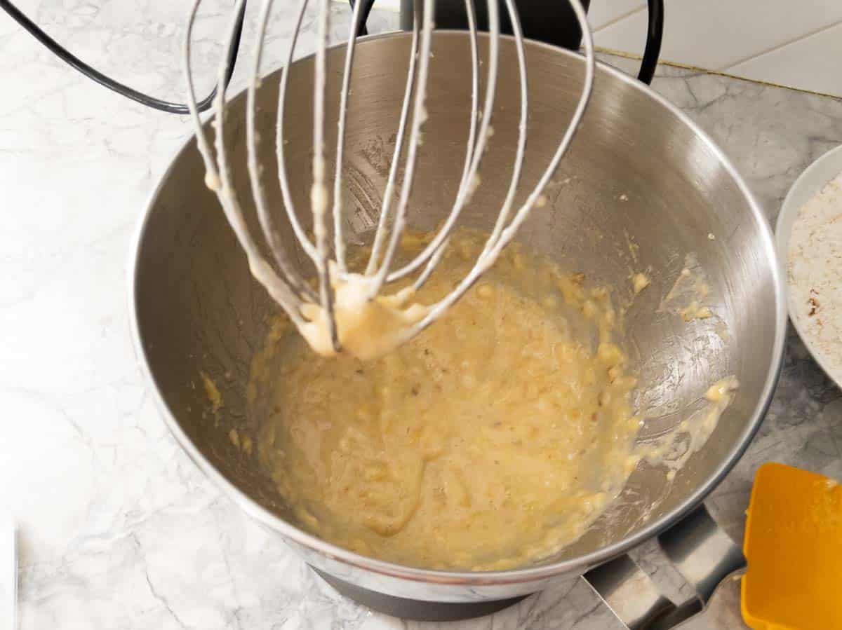 A metal mixing bowl with a whisk attachment contains partially mixed batter, sitting on a marble countertop next to a bowl of dry ingredients and a spatula.
