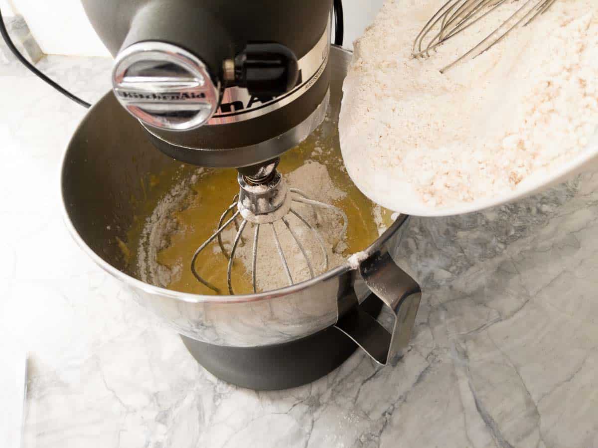 A stand mixer is blending wet ingredients while dry ingredients are being poured in from a white bowl on a marble countertop.