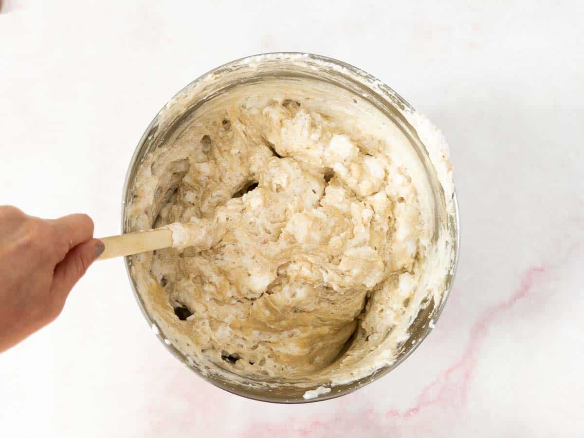 Hand mixing dough with a wooden spoon in a metal bowl on a white surface.