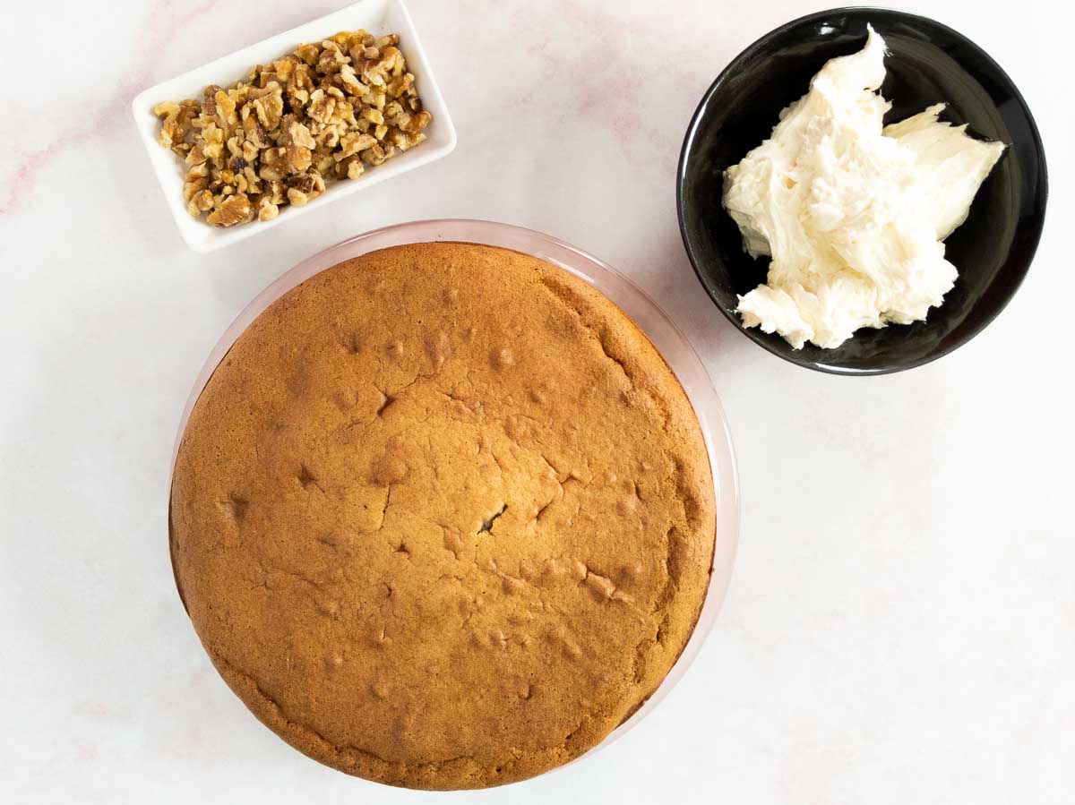 A round, unfrosted cake on a plate, with a bowl of white frosting and a small dish of chopped walnuts on a white surface.