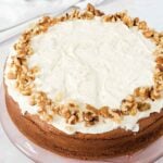 A round cake with white frosting and a border of chopped walnuts on top, displayed on a glass plate.