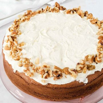 A round cake with white frosting and a border of chopped walnuts on top, displayed on a glass plate.