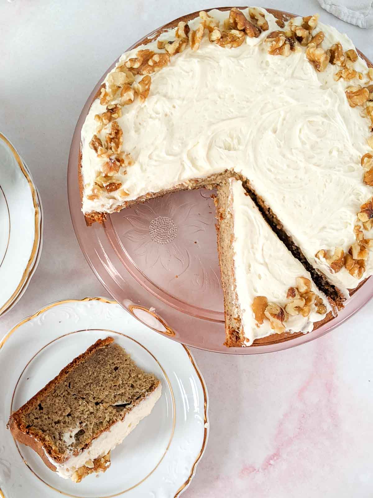 A round frosted cake with walnuts on top sits on a glass cake stand, with one slice served on a white plate nearby.