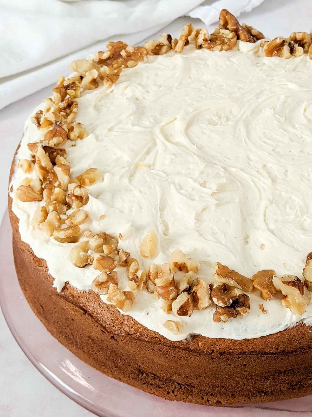 A round cake with white frosting, topped with a ring of chopped walnuts around the edge, sits on a glass plate.
