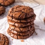 A stack of five chocolate cookies on crumpled parchment paper, with more cookies and a cooling rack in the background.