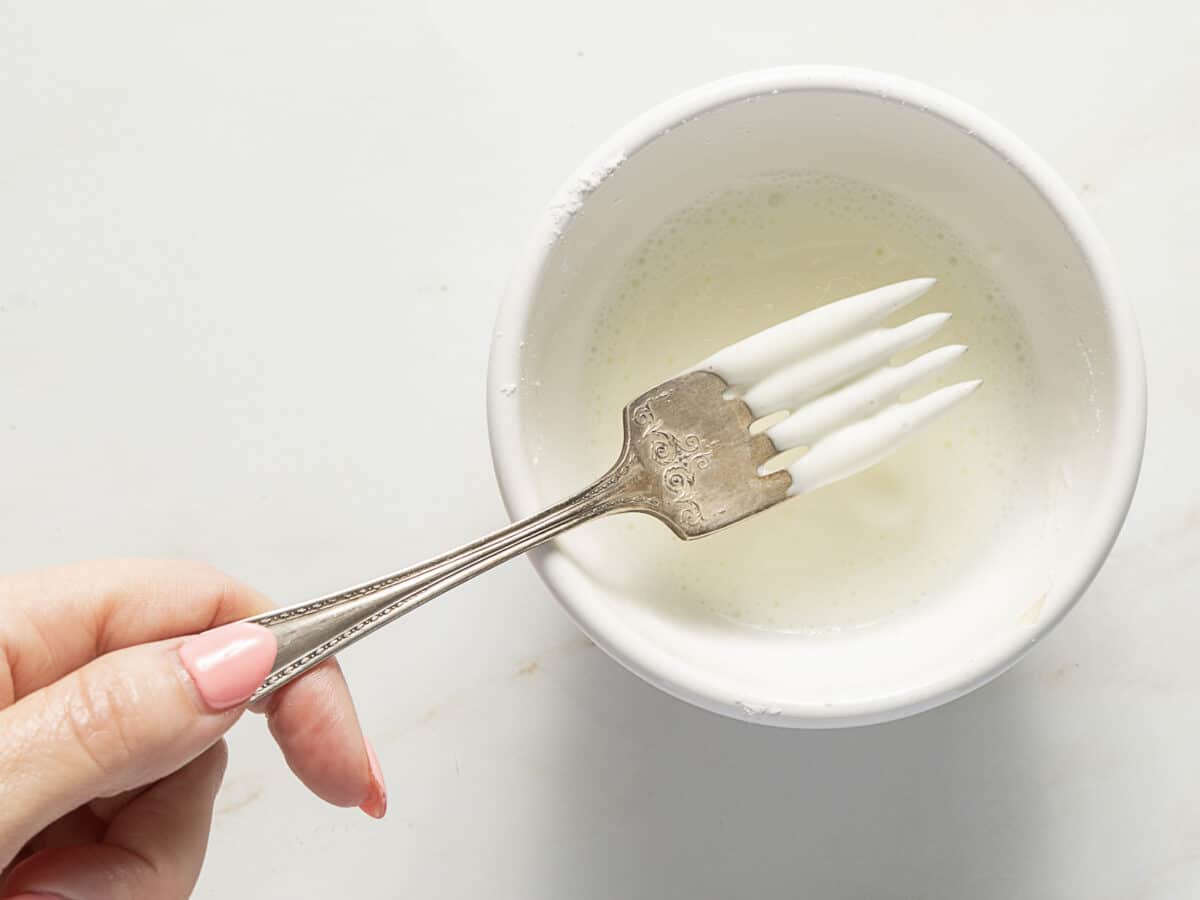 A hand holds a fork coated in white liquid above a bowl filled with the same substance, possibly icing or glaze.