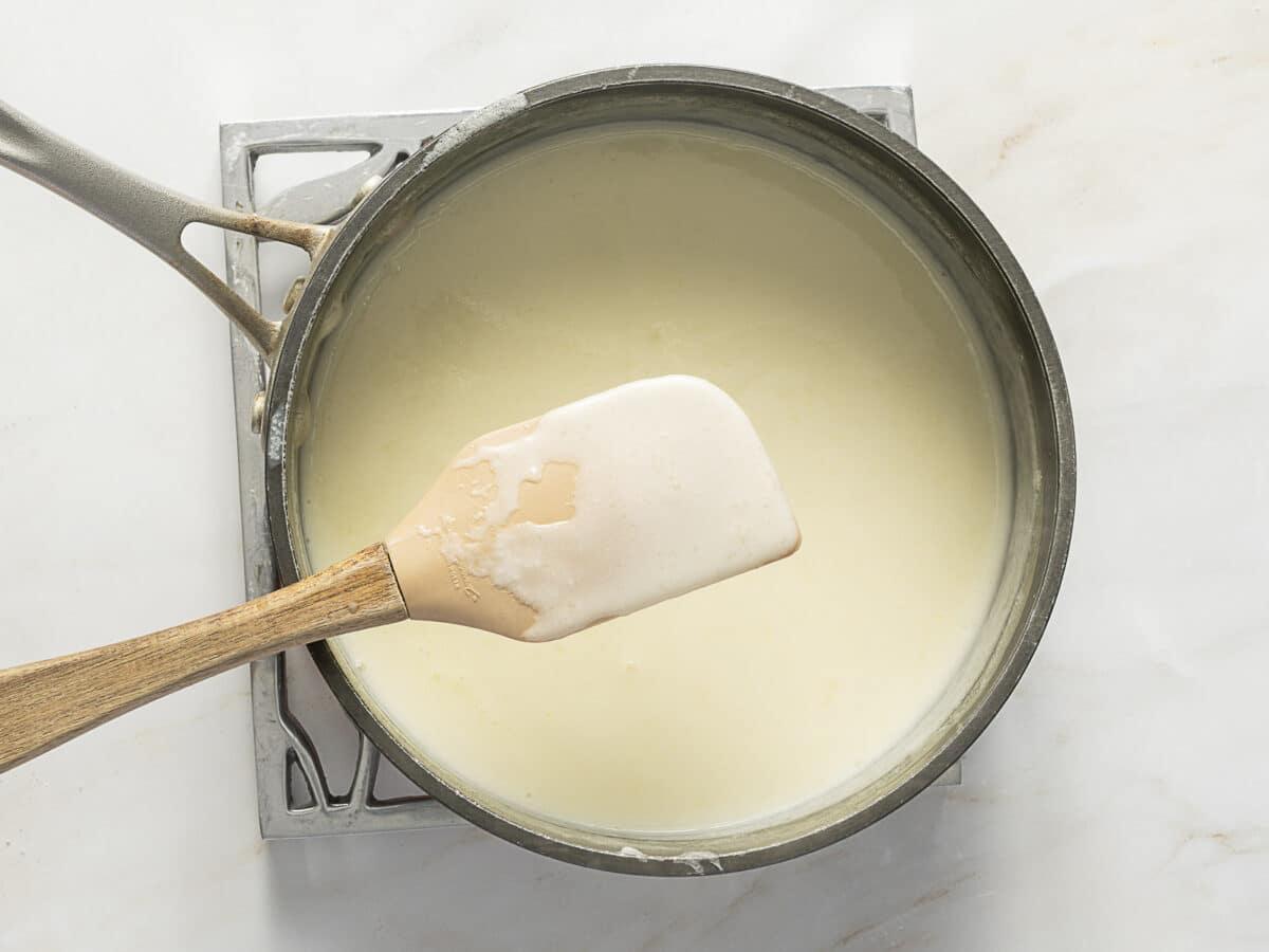 A saucepan filled with creamy white sauce sits on a stove with a wooden spatula resting on its edge, coated in the sauce.