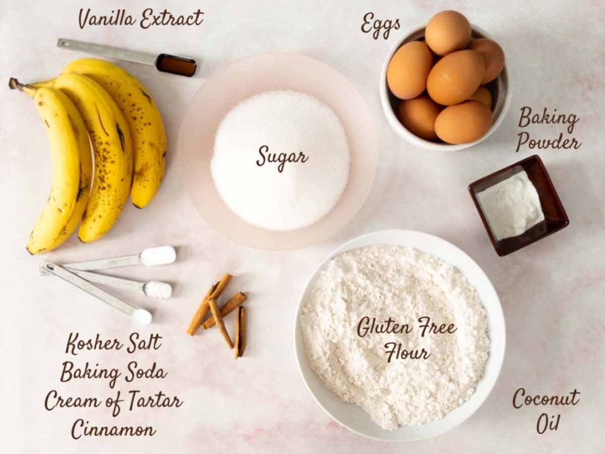 Ingredients for gluten free banana cake are arranged on a white surface, including bananas, eggs, sugar, gluten-free flour, coconut oil, vanilla, baking powder, and spices.