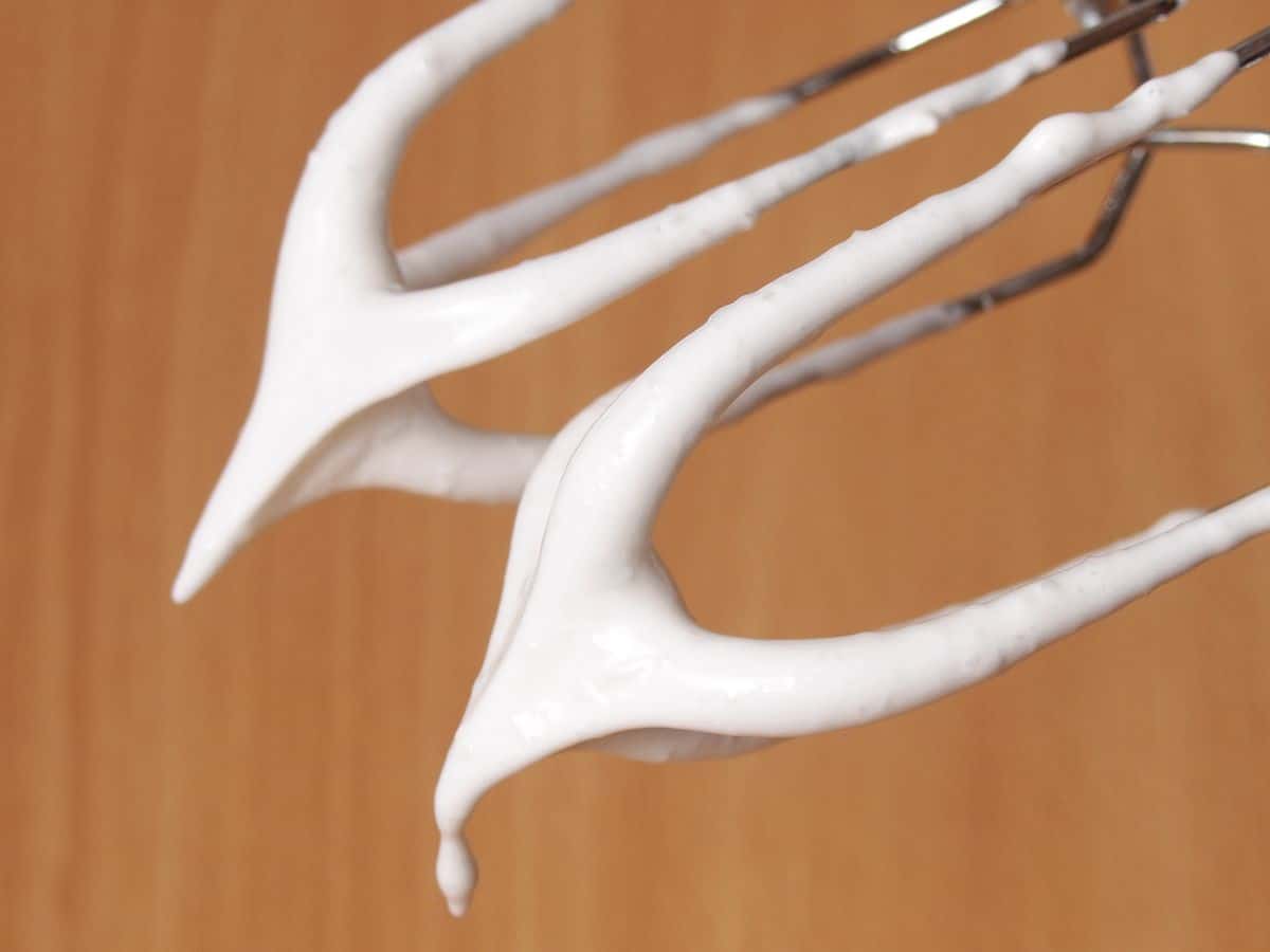 Two metal beaters are coated with stiffly whipped egg whites against a plain wooden background.