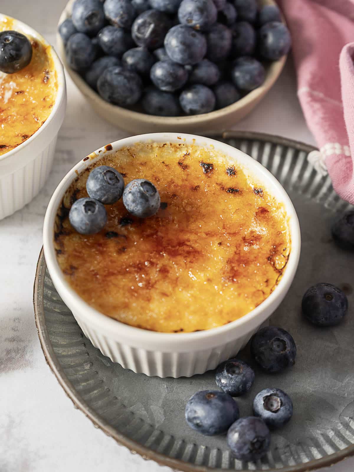 A ramekin of crème brûlée topped with three blueberries sits on a plate, with a bowl of blueberries and a pink cloth nearby.